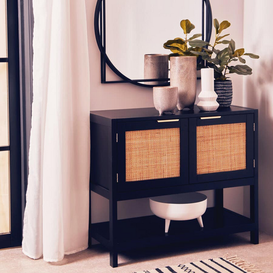 Black console table with wicker doors and decorative items in a room with a mirror and plants.