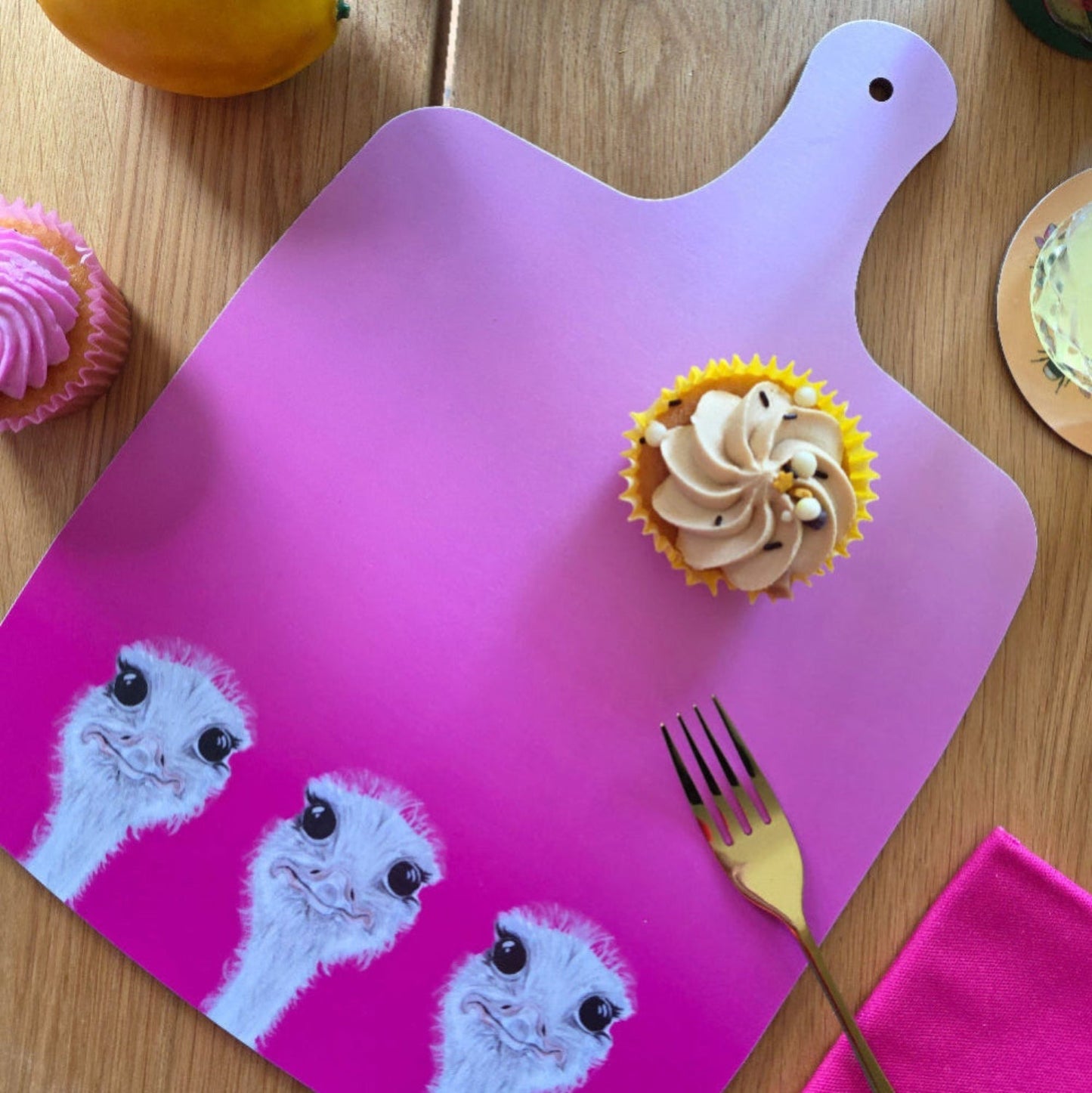 Cupcake on a pink cutting board with ostrich pattern, surrounded by other cupcakes and a fork.