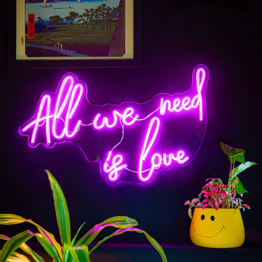 Neon sign with 'All we need is love' text in a room with plants and a framed picture.