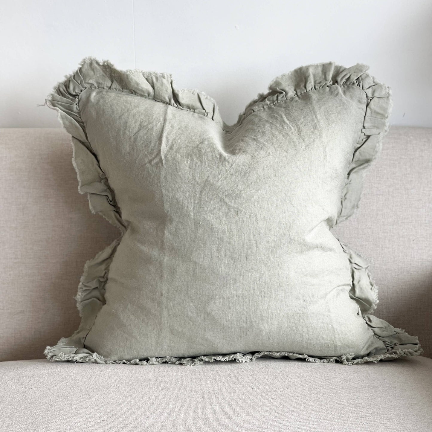 A dusty sage green linen cushion with ruffled edges on a neutral sofa.