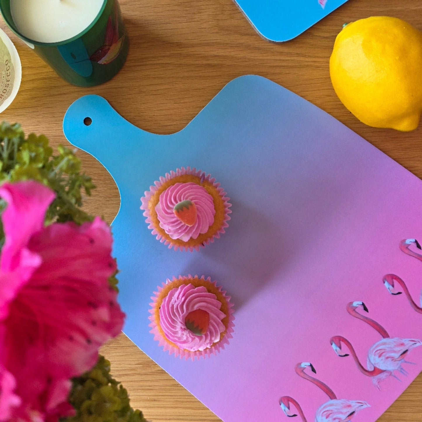 Two cupcakes on a pink cutting board with flamingo designs, surrounded by flowers and a candle.