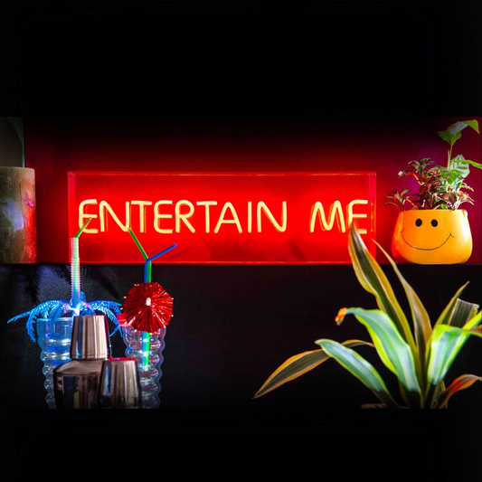 Neon sign that says 'ENTERTAIN ME' with drinks and plants on a dark background