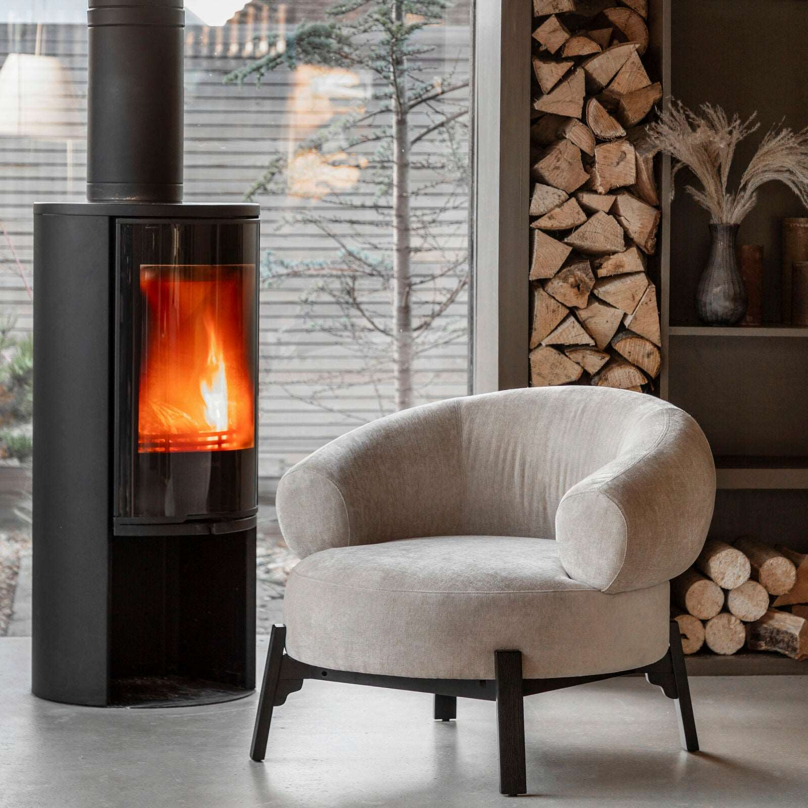 Modern armchair in a room with a lit stove and stacked firewood.