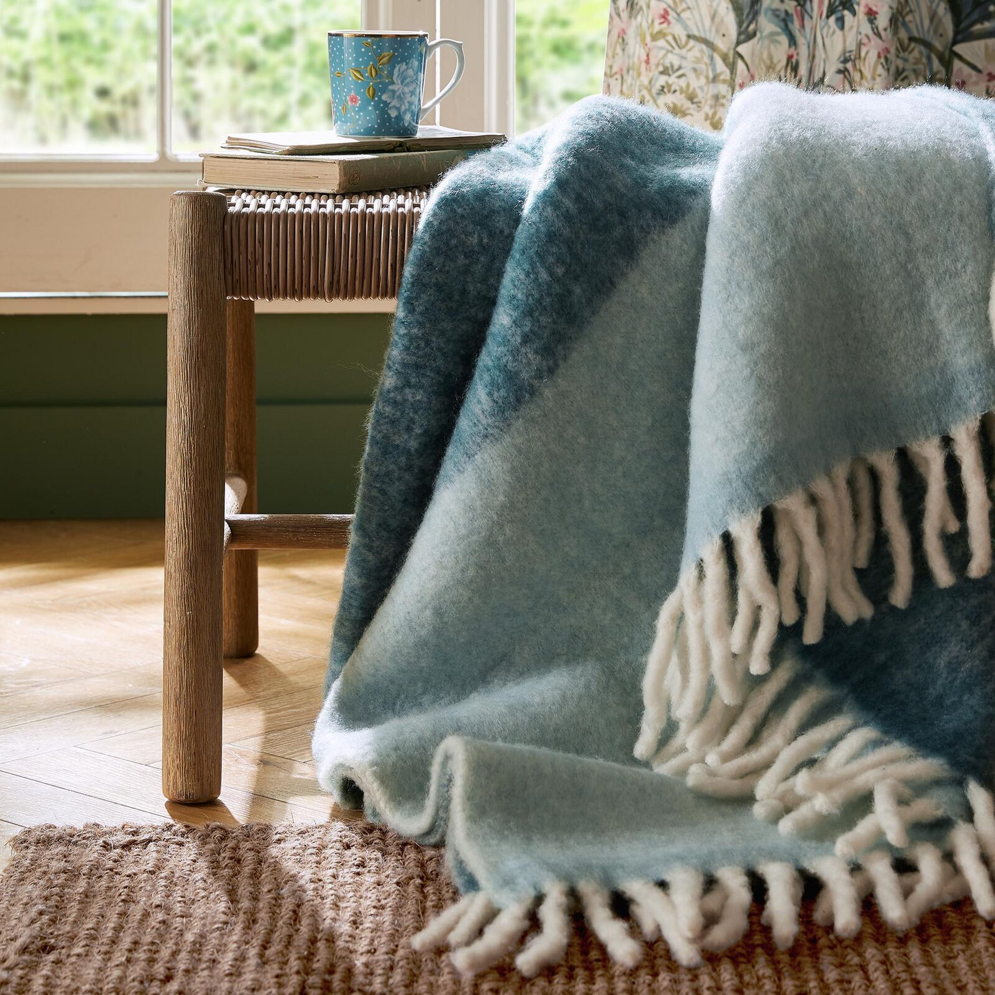 Light blue throw blanket with white fringe draped over a chair next to a wooden table with a mug.