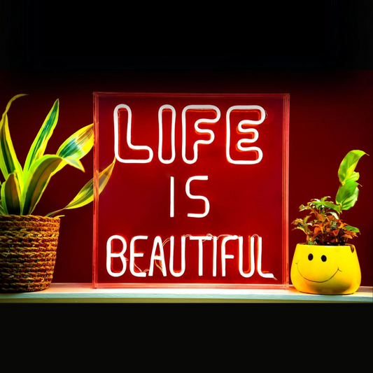 Neon sign with 'LIFE IS BEAUTIFUL' text between two potted plants on a dark background