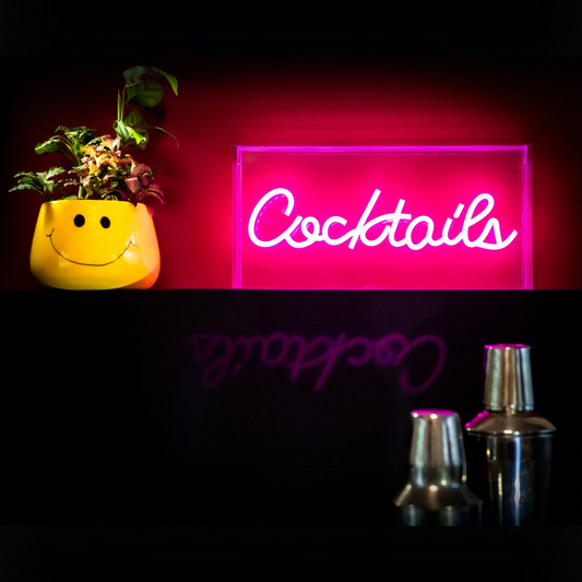 Neon sign with 'Cocktails' text, yellow smiley face planter, and silver shakers on a dark background