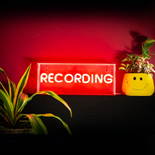 Neon sign with 'RECORDING' text on a dark background with plants.