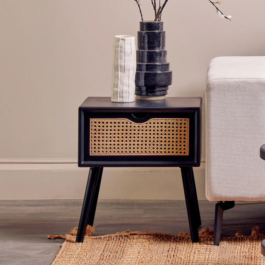 A modern black side table with tapered legs and a woven cane front, styled in a living room next to a light-colored sofa with a dark ceramic vase and a white textured vase on top.