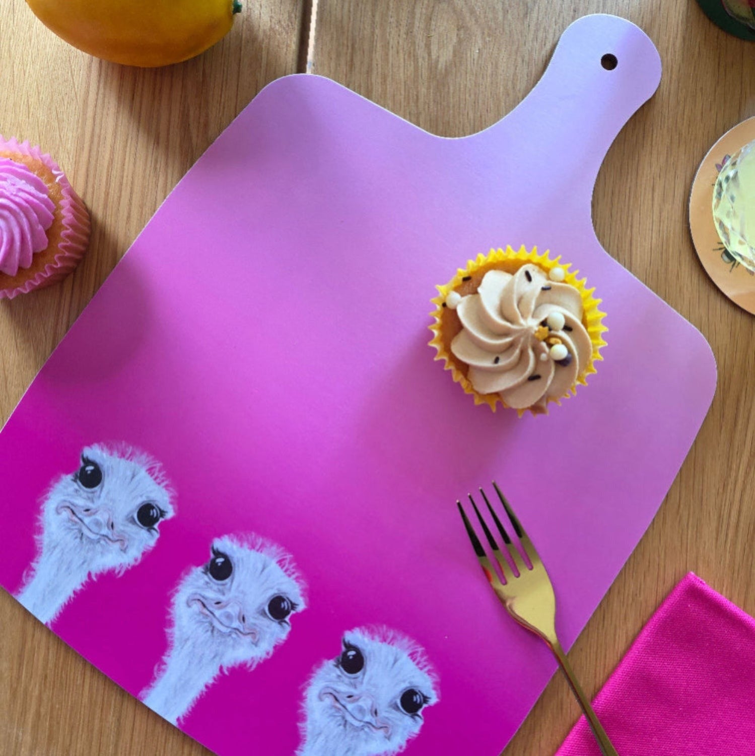 Cupcake on a pink cutting board with ostrich pattern, surrounded by other cupcakes and a fork.