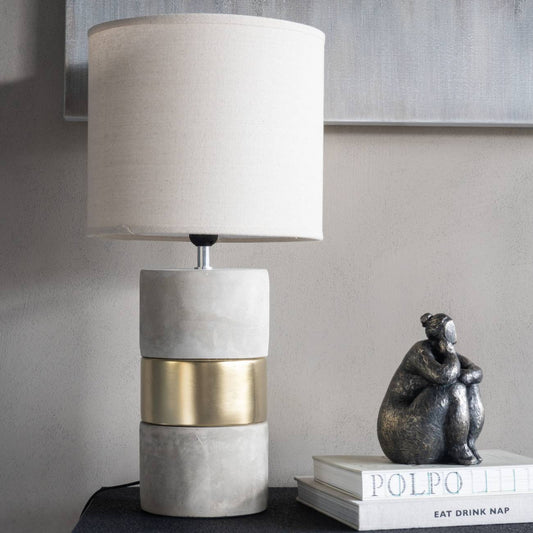 Table lamp with a concrete base and gold accent next to a decorative statue on a shelf.