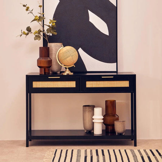 A black console table with two drawers and a lower shelf, styled with vases, a small globe, and a large black-and-white abstract wall art piece above it, set against a warm beige wall.