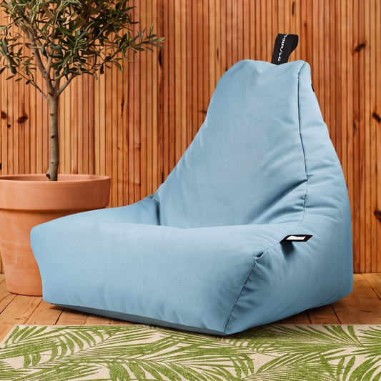 A light blue bean bag chair with a black adjustable headrest and a small black side tag, set against a rustic wood-paneled wall with a potted olive tree and a green palm-patterned rug.
