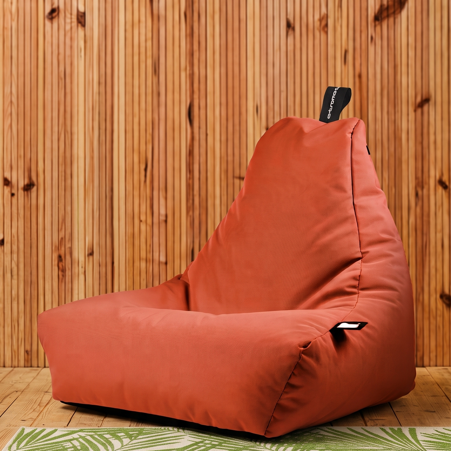A terracotta-colored bean bag chair with a black remote control resting on top, set against a vertical wood-paneled wall and a green palm-patterned rug.