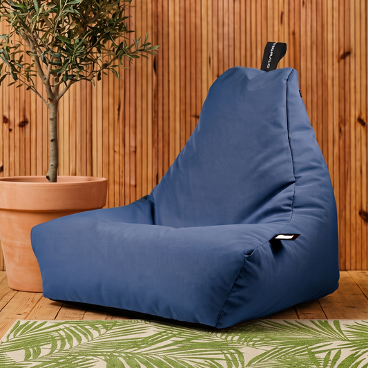 A blue beanbag chair placed on a green and white palm-patterned rug in front of a wooden wall, with a potted olive tree to the left.