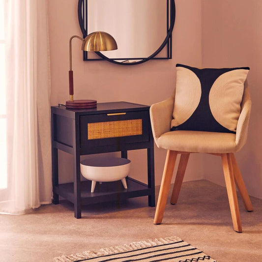A styled interior corner featuring a black-framed oval wall mirror, a dark wood side table with a brass lamp and woven drawer detail, and a beige upholstered armchair with a black and beige accent pillow against a dusty pink wall.