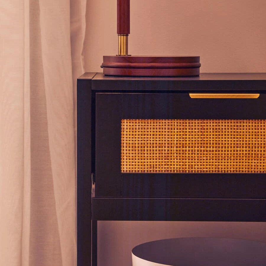 A close-up of a dark wood cabinet with a woven cane drawer front and a gold pull handle, next to a white stool and a lamp base, in a warm beige room setting.