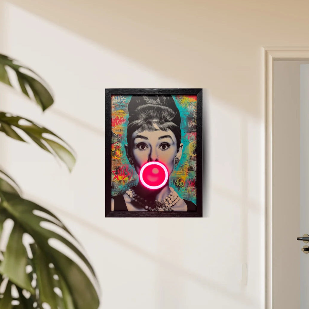 A framed pop-art portrait print of Audrey Hepburn hanging on a beige wall in a room, with a plant leaf visible on the left and a white door on the right.