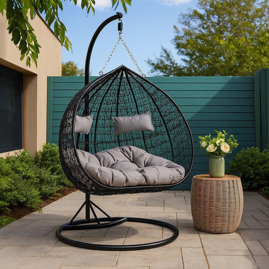 Black egg-shaped hanging chair with gray cushions on a patio, next to a small table with flowers.