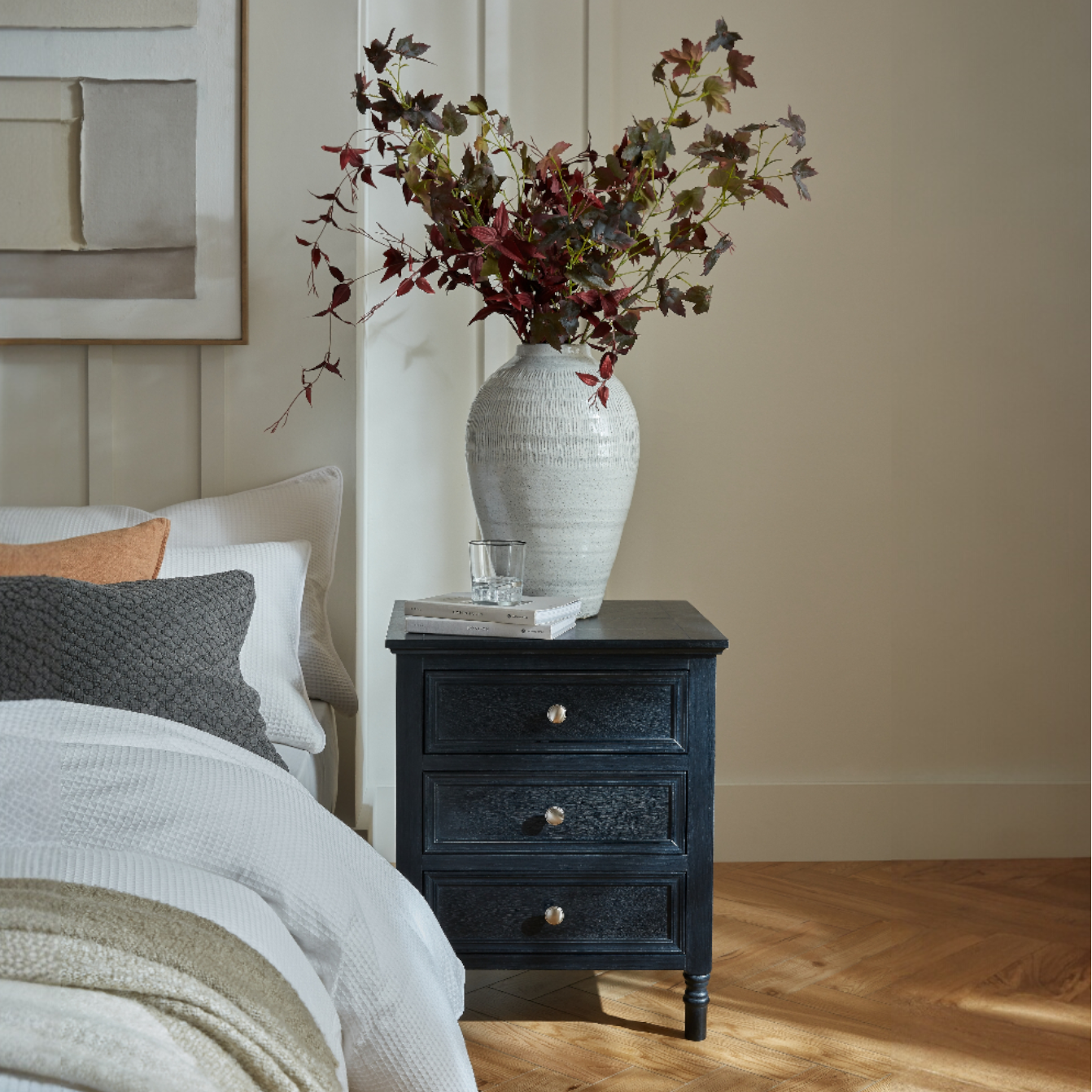 Black nightstand with white vase and floral arrangement next to a bed with pillows and blankets.