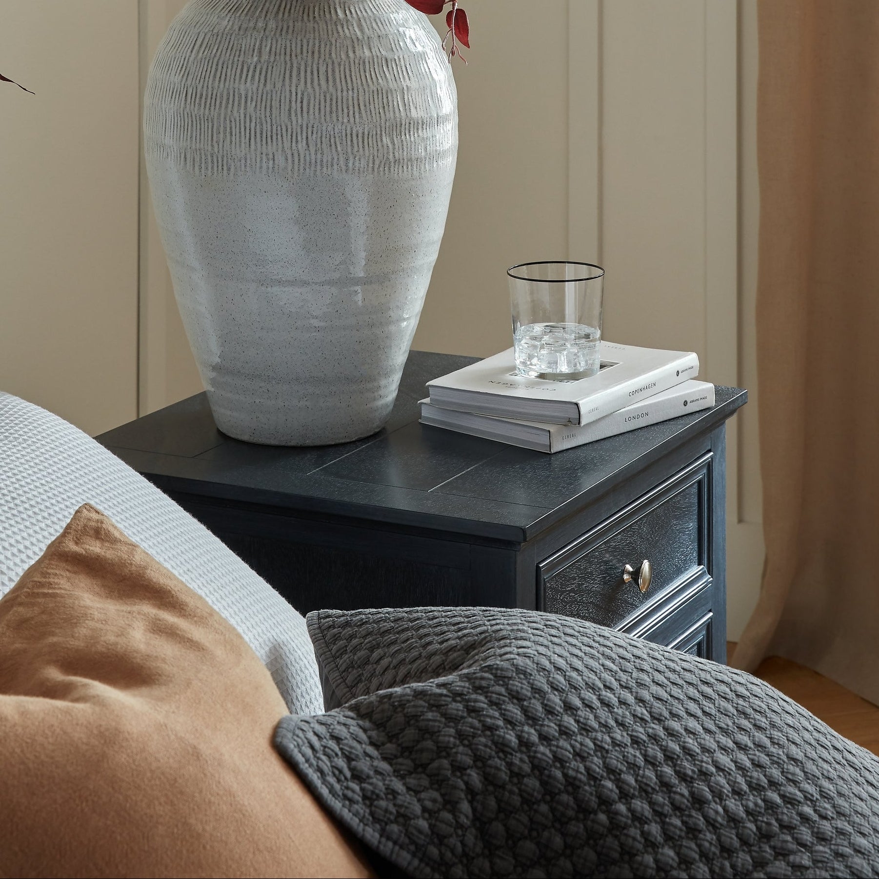 White textured vase with dried flowers on a dark wooden side table against a beige wall.