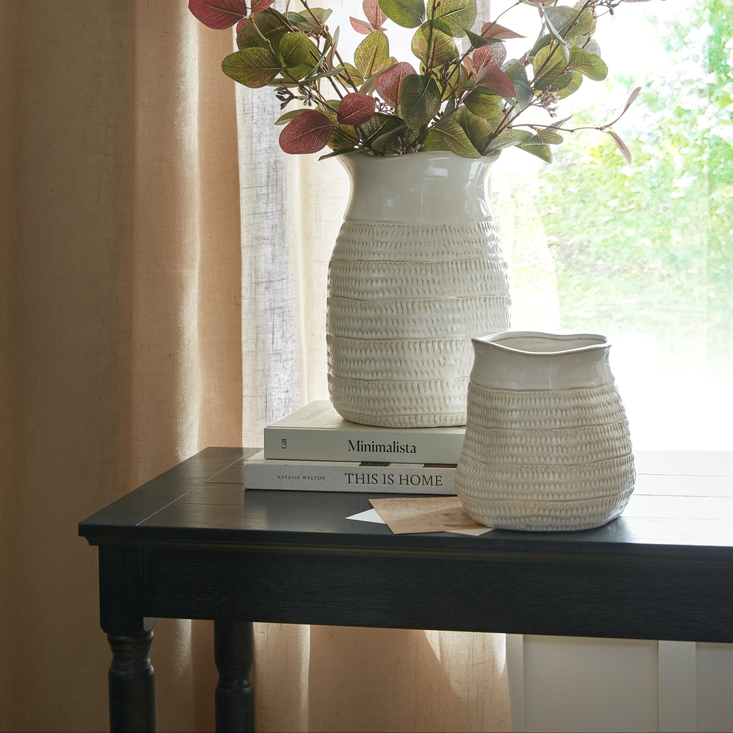 Two textured white vases on a dark wooden table with books and a plant.