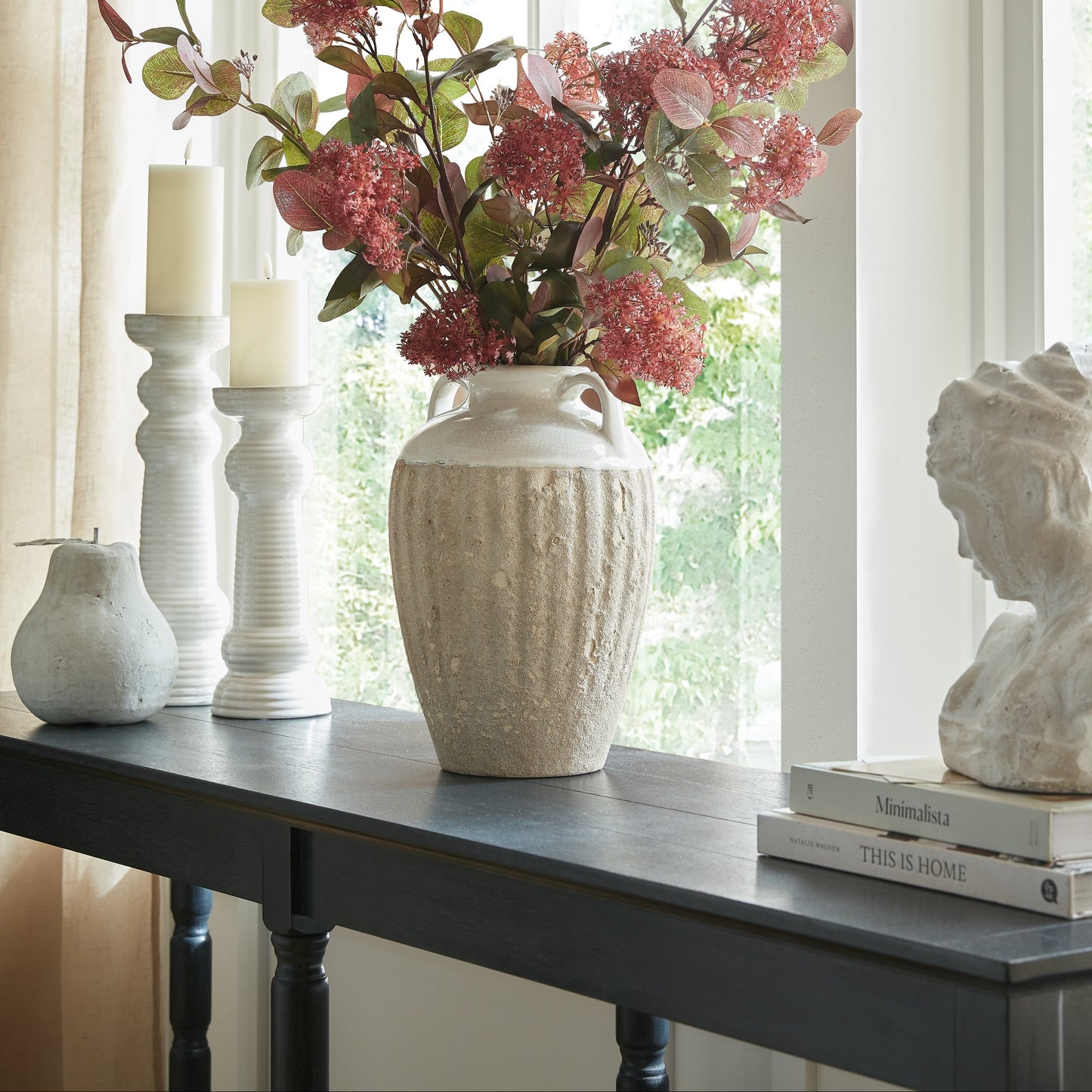 Decorative table setting with a textured vase, pink flowers, candles, and a bust.