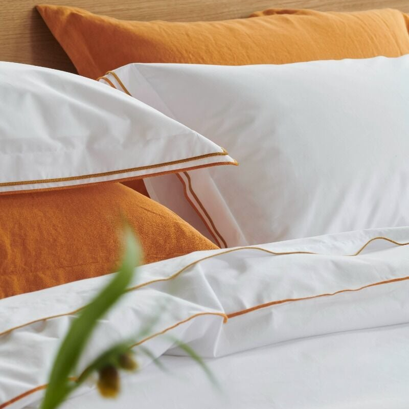 White bedding with gold trim on a bed with orange pillows.