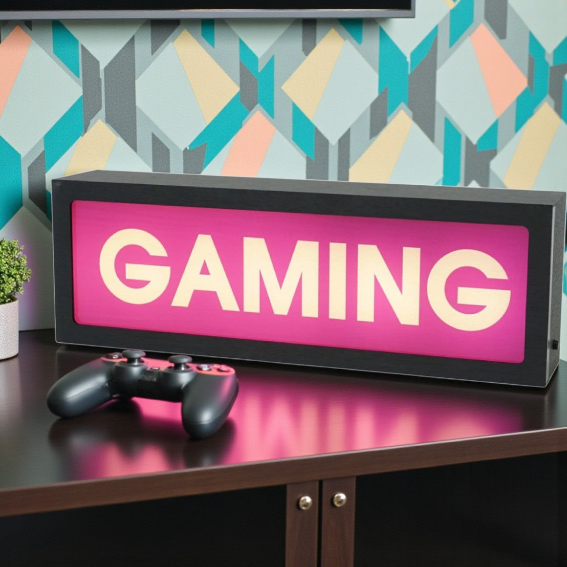 Gaming-themed light box with 'GAMING' text on a desk with a controller, against a geometric-patterned wall.