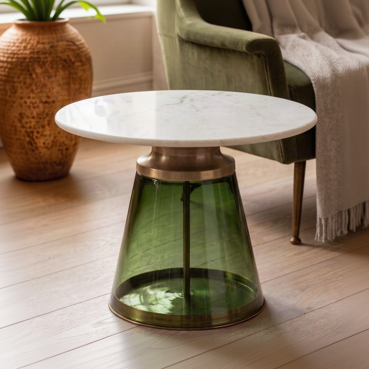 A modern side table with a white marble top and a translucent green glass conical base with brass metal accents, set in a cozy living room next to a green velvet armchair.