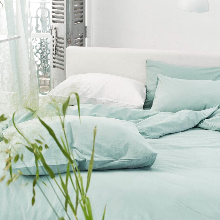 Light green bedding on a bed with white pillows, surrounded by green plants.
