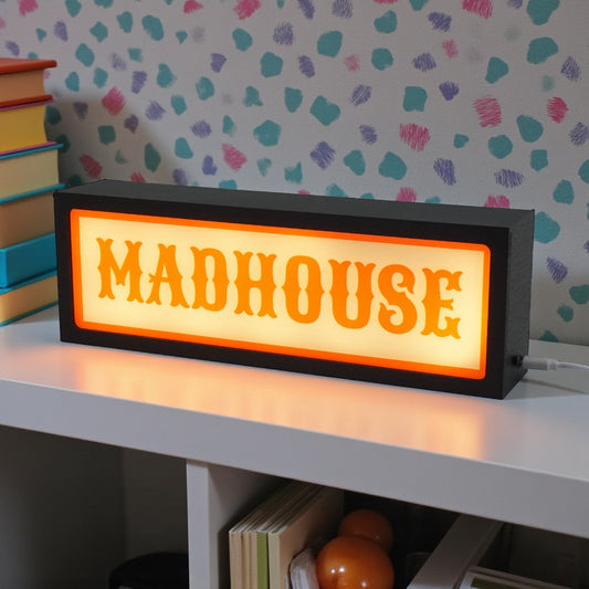 Rectangular light-up sign with 'MADHOUSE' text on a shelf against a patterned wall.