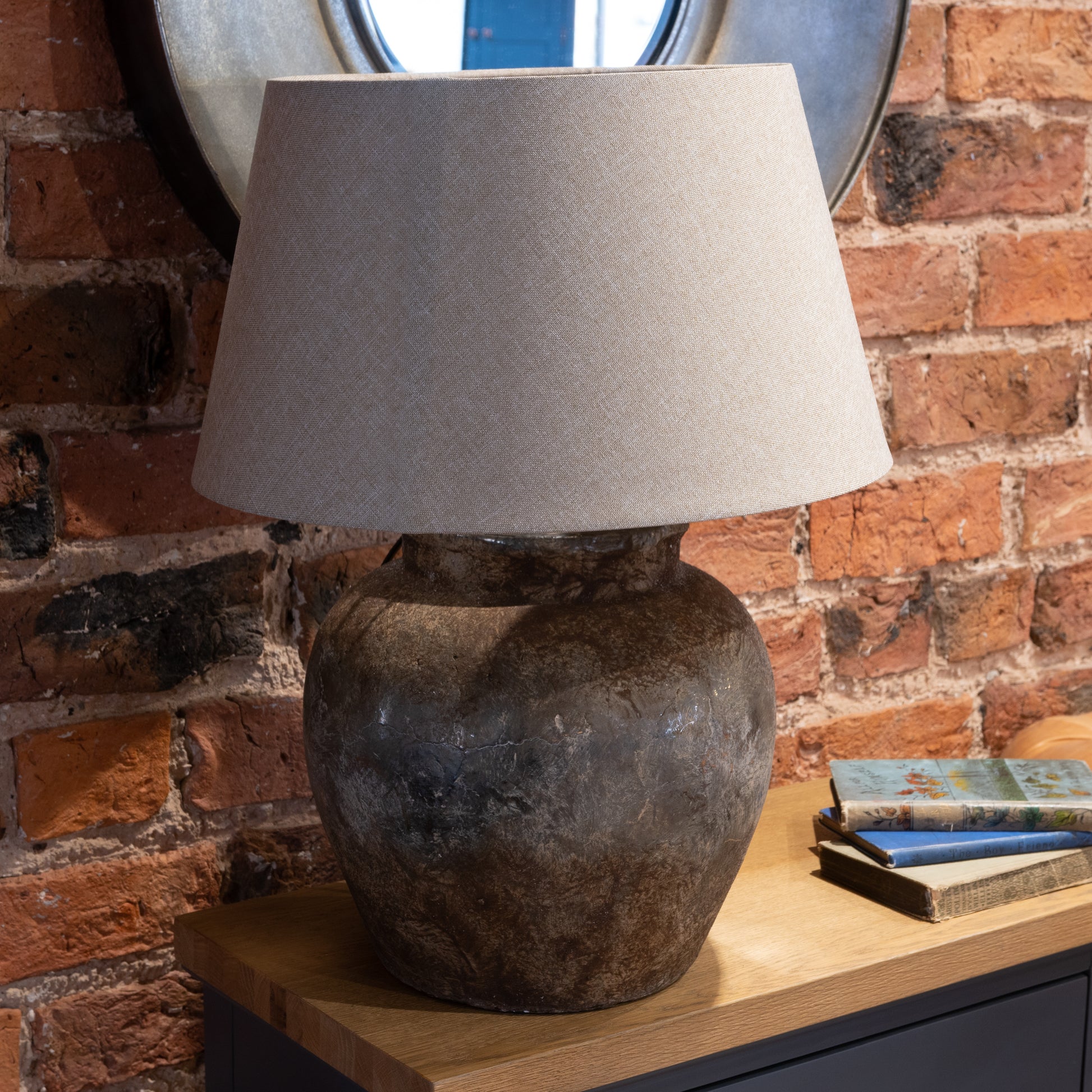 Table lamp with a textured base and gray shade on a wooden surface against a brick wall.