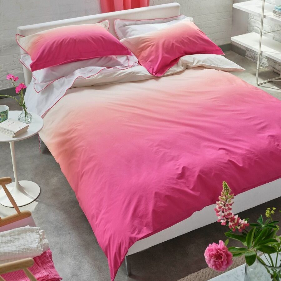 Bed with pink and white gradient bedding in a room with a window and pink curtains.