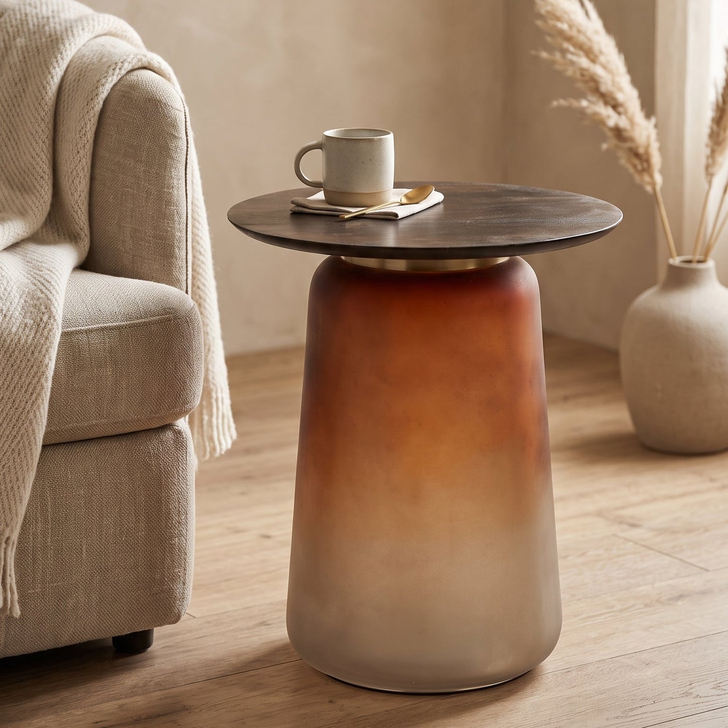 A modern side table with a tapered cylindrical base featuring a warm brown to beige gradient, topped with a round dark wood surface. The table is styled in a cozy living room setting with a mug on top, next to a beige armchair and a vase of dried grass.